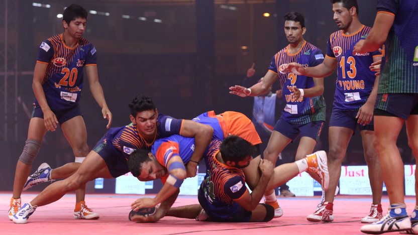 Men grapple in teams on the floor. Three or four men are entangled on the floor with several more stood close by. They are wearing the Indian strip for the Kabaddi team.