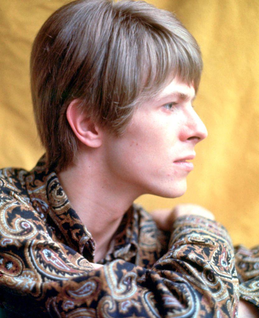 David Bowie, photographed as a young man in side profile. He has sandy hair in a 1960s cut, just longer than his ear. He is wearing a brown and black Paisley pattern shirt.