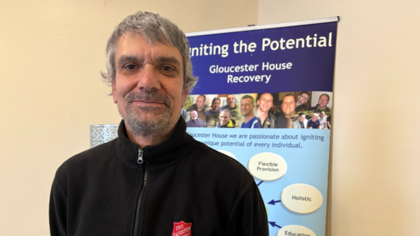 Man in black fleece with a small red logo for Salvation Army in front of a sign which says 'Gloucester House Recovery'