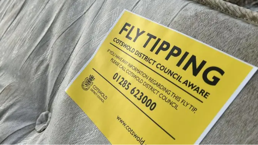 A discarded mattress with a yellow sign saying Fly tipping, Cotswold Disctrict Council aware. And it holds contact information of 01285 623000 to get in touch with the authority if you have any information regarding that waste.