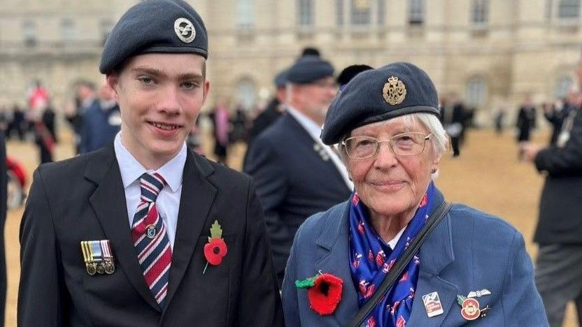 A teenage boy wearing a suit, RAF cap and poppy, stands next to his great-grandmother, who is also wearing an RAF cap, large red poppy, blue coat and blue scarf.