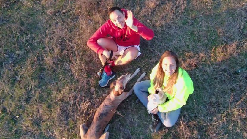 A man and woman waving and looking up at a drone. A dog is lying beside them