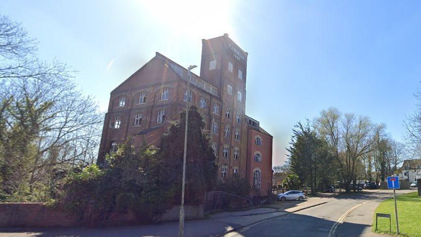 An external view of a large mill building in red brick and will trees and shrubs in front of it. Many of the windows don't have any glass in them and further up the street there are more trees and buildings just visible.