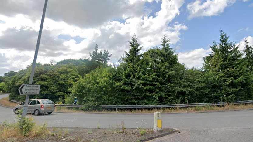 A roundabout with lots of large trees and a sign reading "Town centre, Leisure centre, Chawson" on the left hand side. A Citroen car can be seen driving round.