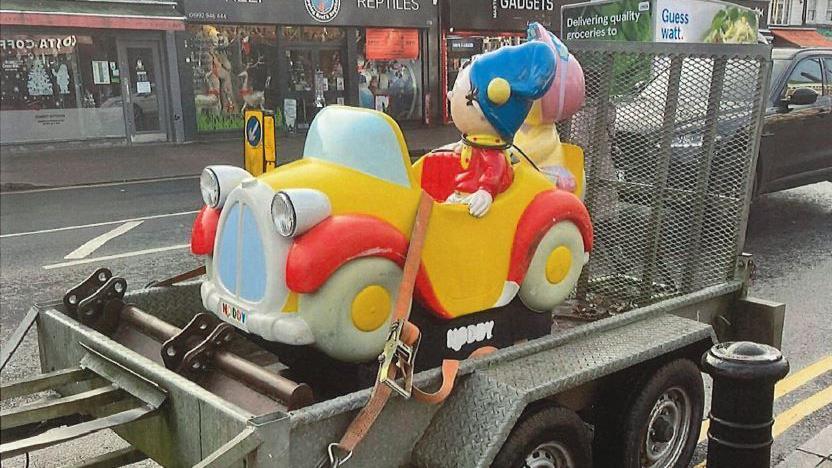 The statue of Noddy is strapped to a luggage cart ready to be towed away. The cart is on the road and a number of shops can be seen behind it. 