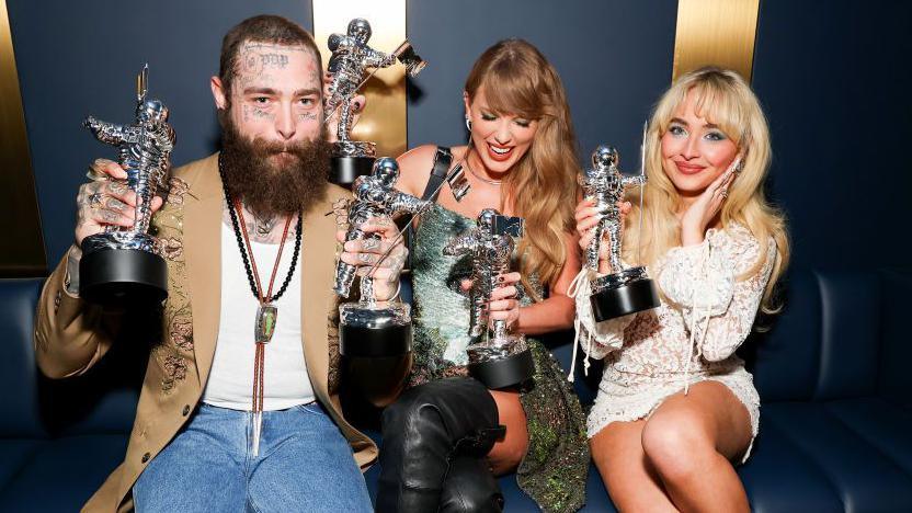 Singers Post Malone, in jeans and a beige jacket, Taylor Swift, in a silver dress, and Sabrina Carpenter, in a white dress, hold their astronaut shaped VMA trophies and smile