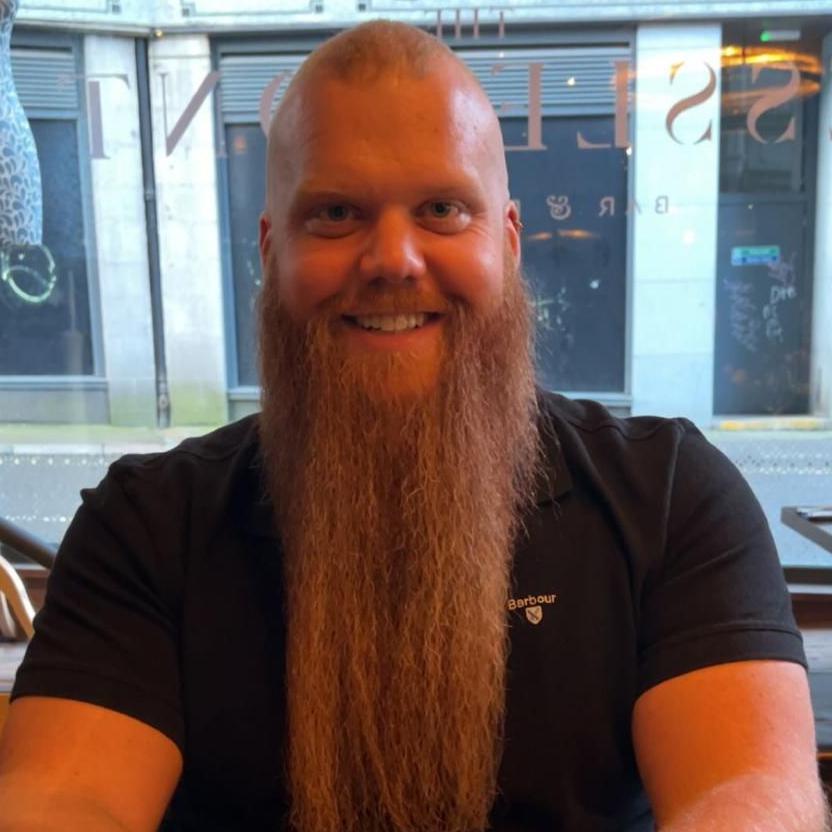 A man with a very long, straight bear and a shaved head is smiling at the camera. He is wearing a black T-shirt and sitting in front of a window.