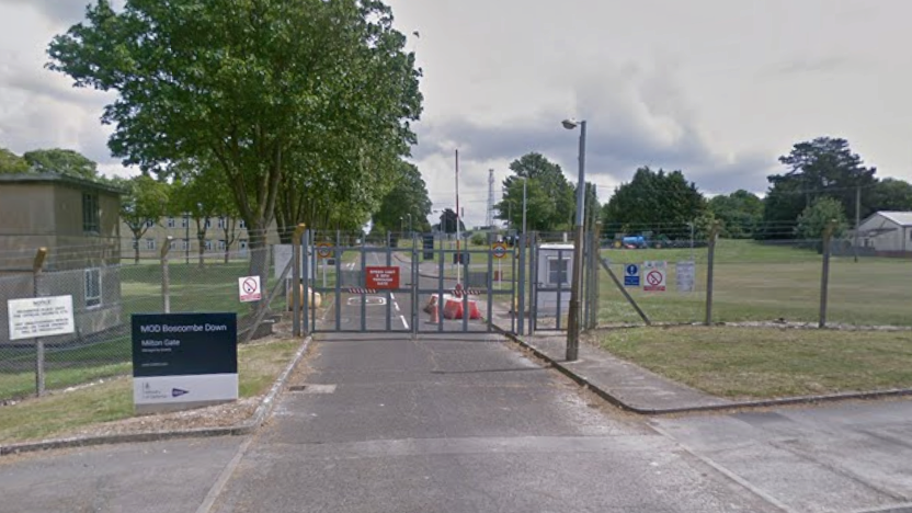 The gate to MoD Boscombe Down. The airfield can be seen in the background, including some hangars and a large tree.