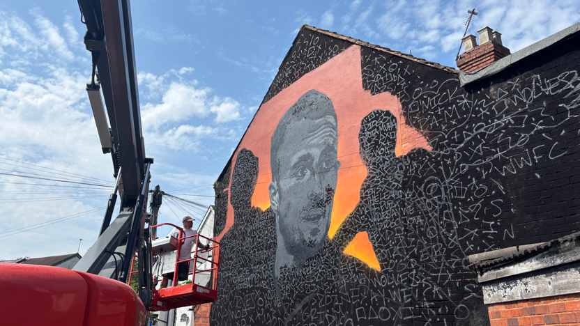 A man painting a black and orange mural.  We can see a close-up of the mural in black with white letters on the painting which makes out a man's face on the wall of a building.
