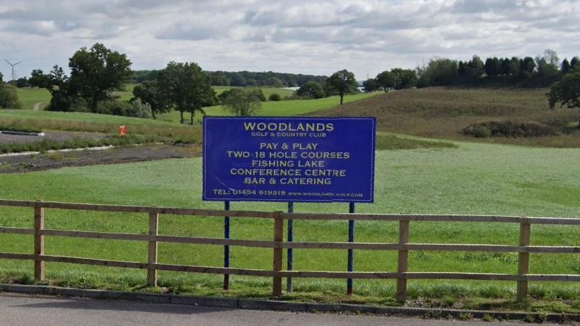A blue sign saying Woodlands Golf and Country Club, above a fence with green grass and trees behind.