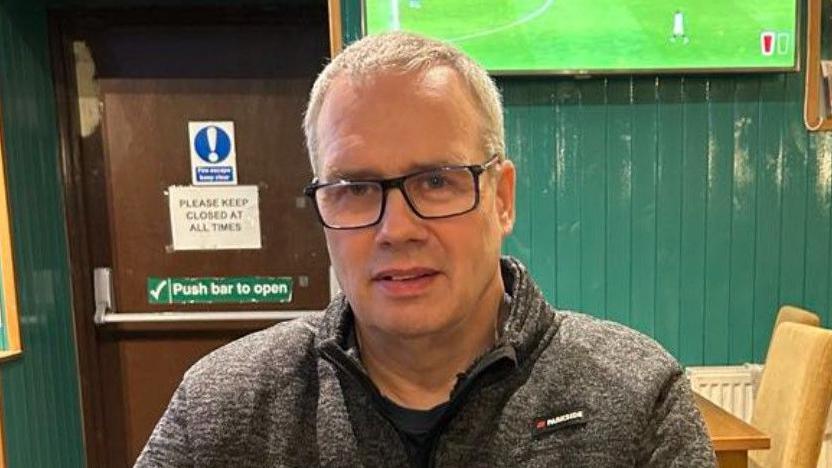 Donald Mac Isaac sitting in a bar or restaurant.  He has short grey hair and black rimmed glasses.  He is wearing a grey fleece jacket over a black t-shirt.