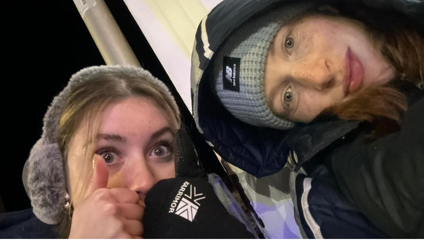 A selfie of two women, one wearing ear muffs, gloves and giving a thumbs up, and the other wearing a hat and a coat with her hood up. The photo is taken at night and there's a lamppost behind them.