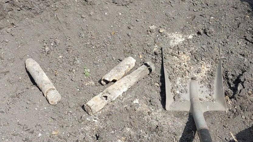 Three unexploded bombs among soil and rubble, they are white in colour and appear corroded. A shovel can be seen on the right hand side of them.