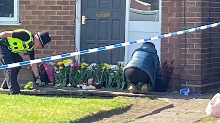 A woman kneeling down by the house and placing flowers by the door. A police officer stands to the left of the house.