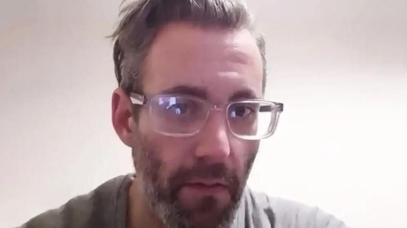 A picture of Michael Wheeler. He is wearing clear framed glasses and has grey hair combed backwards and a beard. He is wearing a grey t-shirt and is sitting against a plain background.