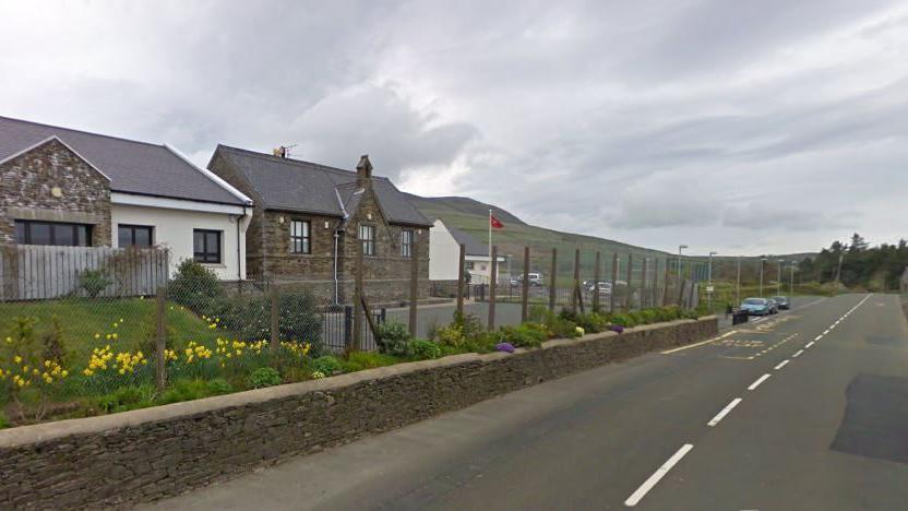 Three stone buildings in a line to the left with grass and tarmac in front of them. To the right is a straight main road. It is a cloudy day.