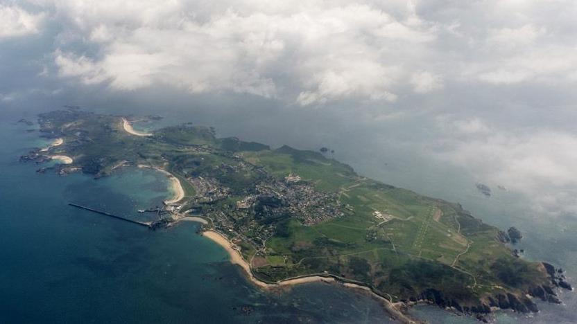 A shot from above of Alderney. Green land is visible with a beach and blue water surrounding it.