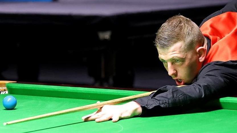 David Church playing snooker while wearing a red waistcoat and black shirt. Under a bright light, he is leaning over the green felt of a table, his cue at the ready, concentrating on a shot. 