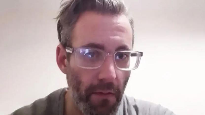 A picture of Michael Wheeler. He is wearing clear framed glasses and has grey hair and a beard, and is wearing a grey t-shirt. 