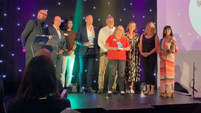 People stand on a stage as they accept the award. The group is made up of five men and four women. They are posing for a picture. They are standing in front of a black backdrop with small illuminated lights.