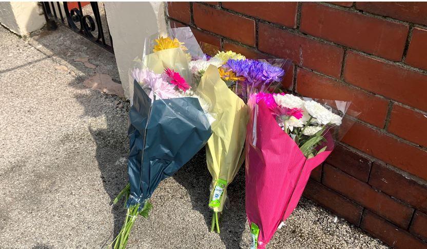 Flowers wrapped in pink, yellow and grey lean up against a red brick wall outside the scene of a fatal house fire