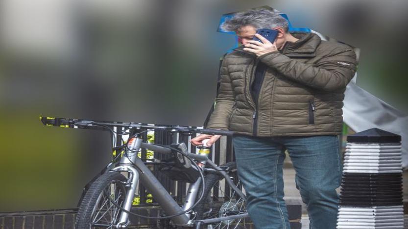 Howard Phillips wearing a brown coat and jeans while on the phone. He is using his left hand to press a USB stick into the seat of a bicycle.