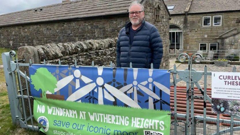 A man stands behind a banner saying 'No windfarm at Wuthering Heights' which is attached to a gate. He wears a blue puffer jacket and black glasses. He is middle aged, white, and has greying wispy hair and a goatee. He stands in the yard of a rural property, possibly a farmhouse.