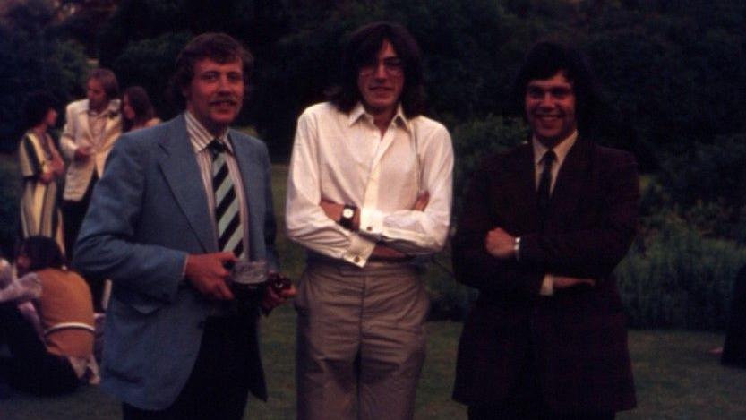 A colour photograph taken in the mid-1970s of three men wearing different coloured suits. Behind them are other people chatting and sitting on the floor outside.