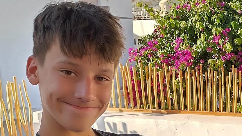 Murray smiling at the camera while sitting in his back garden on a sunny day. He is sitting to the left of the picture, and there are pink flowers and greenery in the background. 