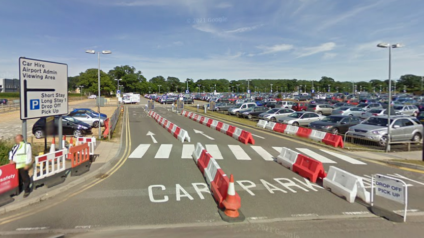 A car park filled with cars. White painted signs on the road read "car park". There are temporary white and orange plastic barriers at the entrance. There is also a sign which reads "Drop off, pick up".