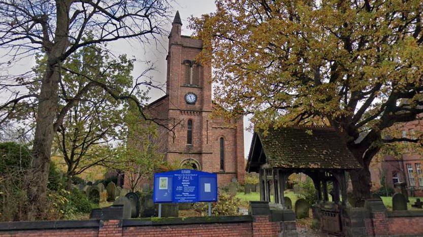 The church tower of St Paul's in Withington seen from a road with trees in the foreground and a blue sign for the building sat on a churchyard wall. 