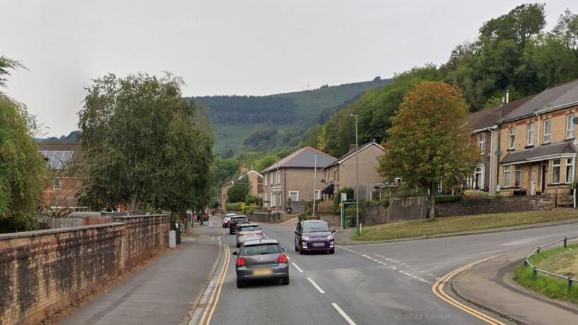 St Mary Street, Risca, with a residential area to the right, cars passing each other on the road, trees visible in front of the houses and forest in the background. 