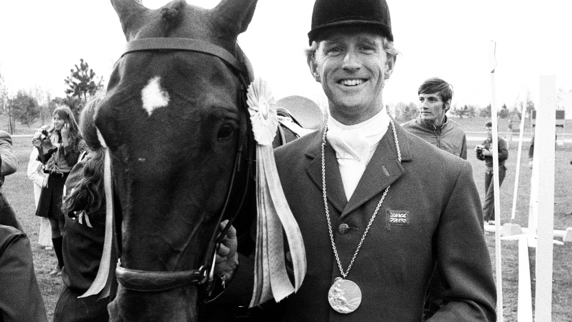 Richard Meade standing with his horse. He is wearing a riding hat and jacket with the British flag sewn into his lapel. He has a medal around his neck. 