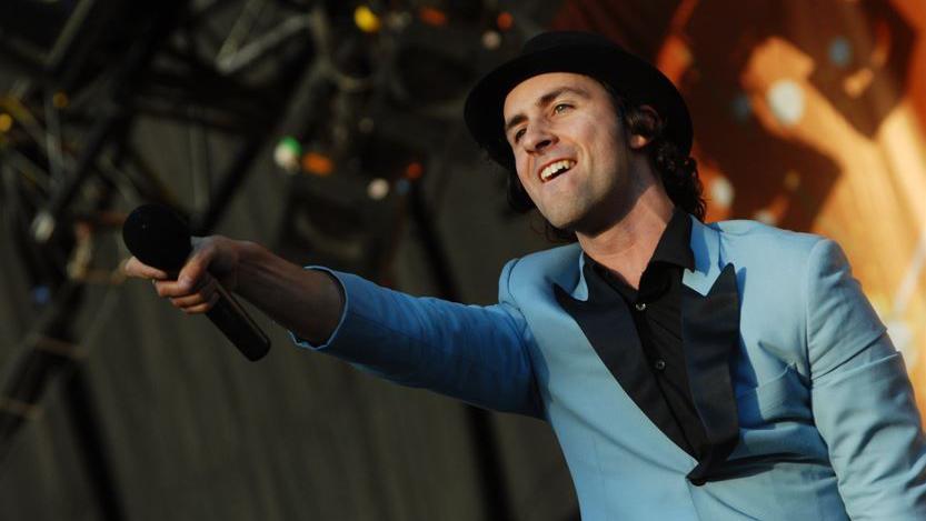 Paul Smith of Maximo Park performs on stage. he is wearing a light blue suit and hat. He is holding his microphone out to the crowd.