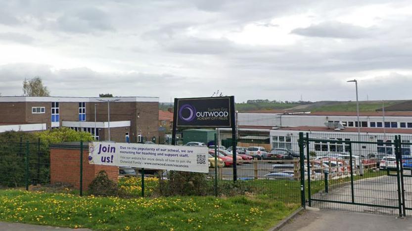 A black sign saying Outwood Academy City Fields sits in front of a low-rise school buildings 