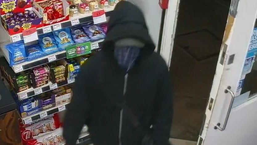 A still image of the offender taken from the store's CCTV. He is wearing a dark coloured hoodie and has a blue piece of material covering his face.