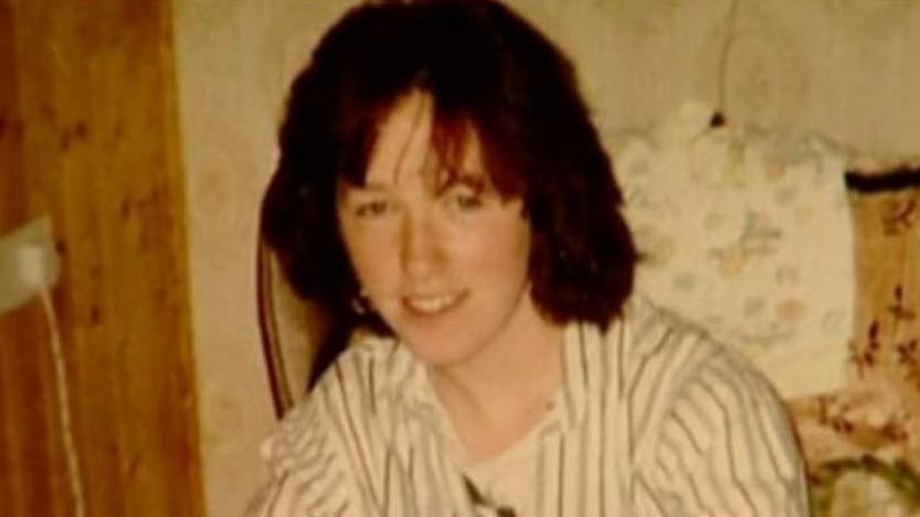 A woman, with brown hair, sitting on an old fashioned couch, smiling at the camera. She is wearing a white shirt with blue stripes on it. 