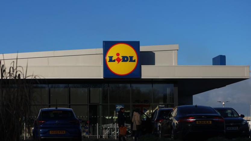 A generic photo of a Lidl supermarket, the building is grey, there are cars in a car park parked out the front. In the middle is the blue, yellow and red Lidl logo. 