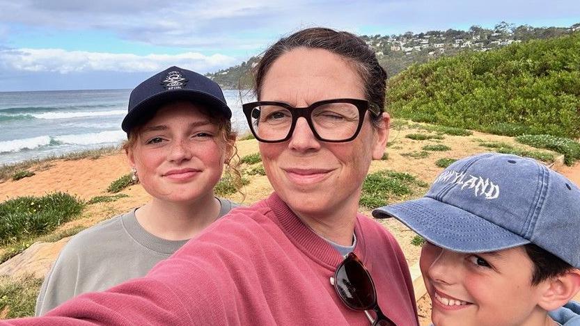 A woman with dark hair tied back. She is wearing glasses and a pink jumper. She is stood next to two smiling children, a boy and girl. They are posing for a selfie picture on a beach.