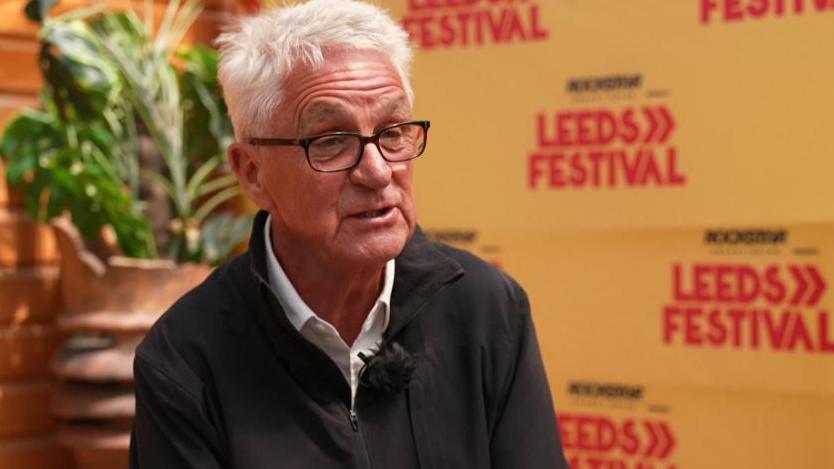 A man with white hair and glasses against a Reading and Leeds festival background.