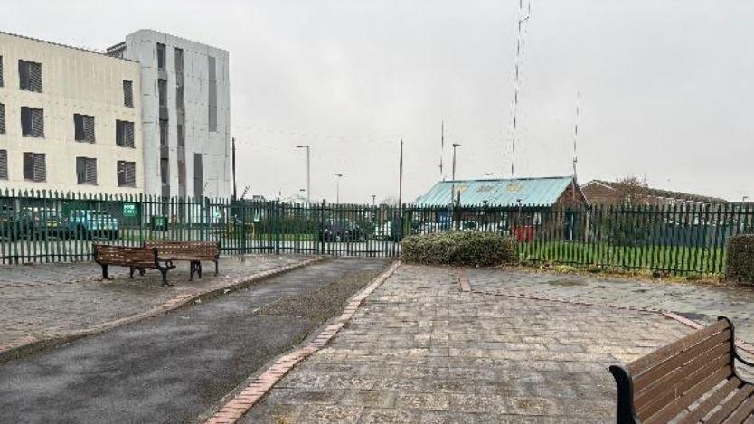 An outdoor paved area with benches and a narrow path lined by red bricks. A green metal fence surrounds the space, separating it from nearby buildings, which include a white structure and a smaller blue-roofed building. The sky is overcast and the pavement is wet.