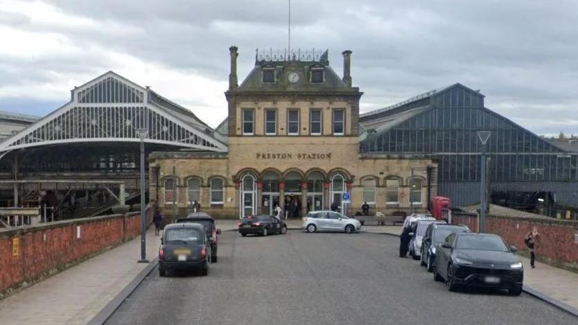 Preston Railway Station front entrance. The building is a light brown brick, and there are taxis and cars parked on either side of the front entrance.