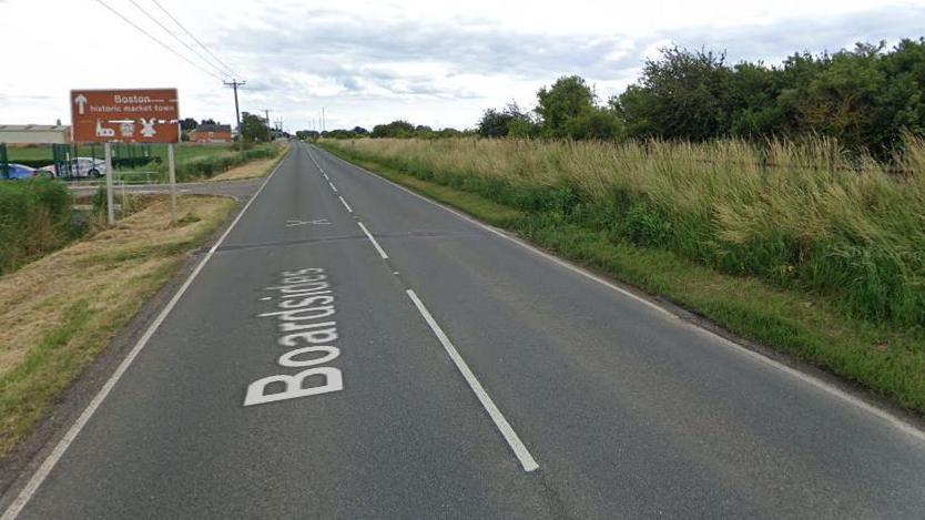 A Google street map image of a road. There is a sign up ahead on the left that reads 'Boston, historic market town' with an arrow next to it pointing ahead. The road is lined either side by grass verges and trees.