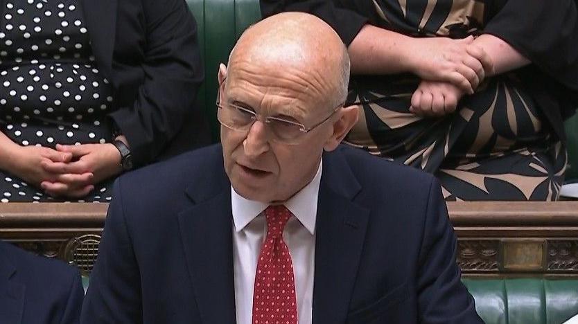 Image shows a man wearing a dark blue suit with a red spotty tie standing in the house of commons. He is wearing glasses. Behind him are fellow MPs sitting on the green benches in the House of Commons. 