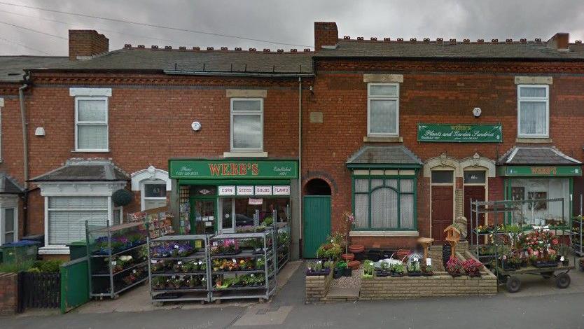 The front of a shop, which has green signage above the door, has racks of flowers outside. It is situated among a row of terraced houses.