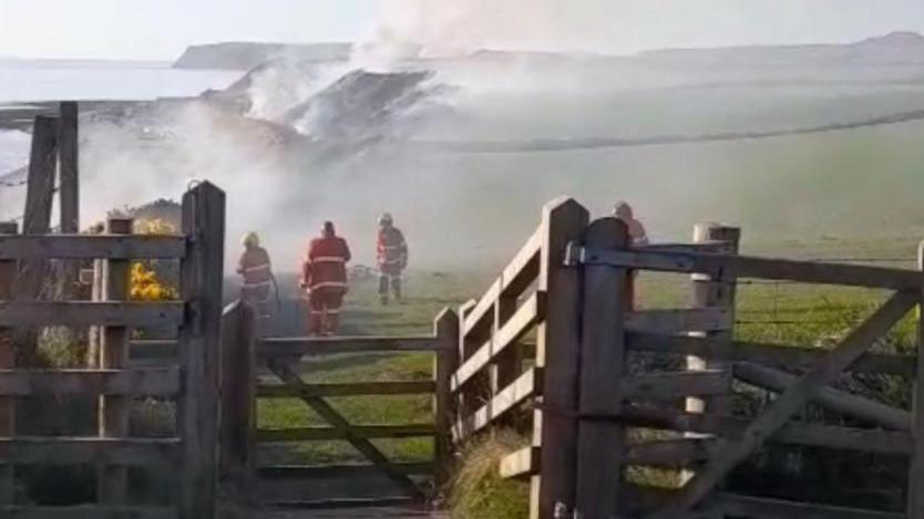 Three firefighters in red uniforms tackle a fire in the countryside behind a fence. Large plumes of smoke can be seen. 