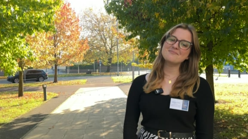 Sienna smiles into the camera. She wears spectacles, has long brown-blonde hair and has a white name card on her black top. 