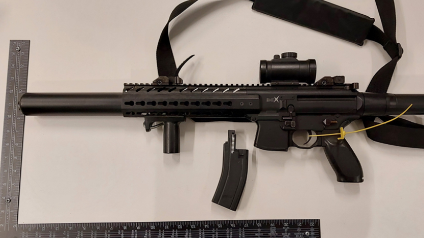 A long barrelled black firearm is placed alongside an L shaped ruler. The firearm's magazine is separate to the body of the weapon, and there is a yellow plastic tag wrapped around the trigger.