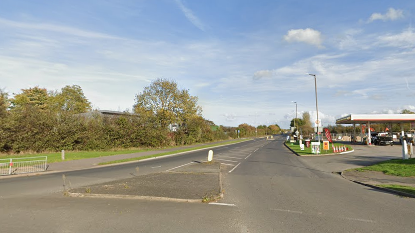 An image of a road taken from a roundabout. There are trees to the left and a petrol station to the right. The sky is blue.