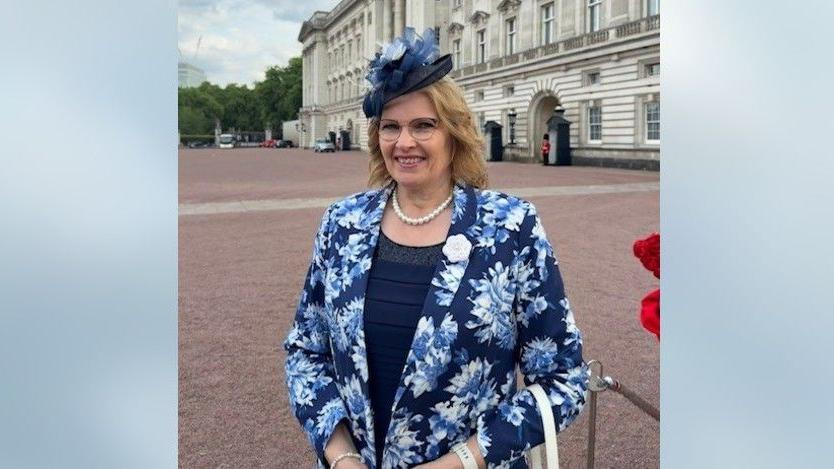 Jilly Crofts in front of Buckingham Palace in a blue, flowery suit jacket and hat